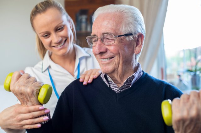 Female Physiotherapist Helping Senior Man To Lift Hand Weights