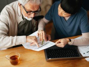 From Above Photo Of A Senior Man Using A Laptop Computer And Paying Bills With Serious Nurse