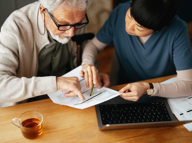 From Above Photo Of A Senior Man Using A Laptop Computer And Paying Bills With Serious Nurse