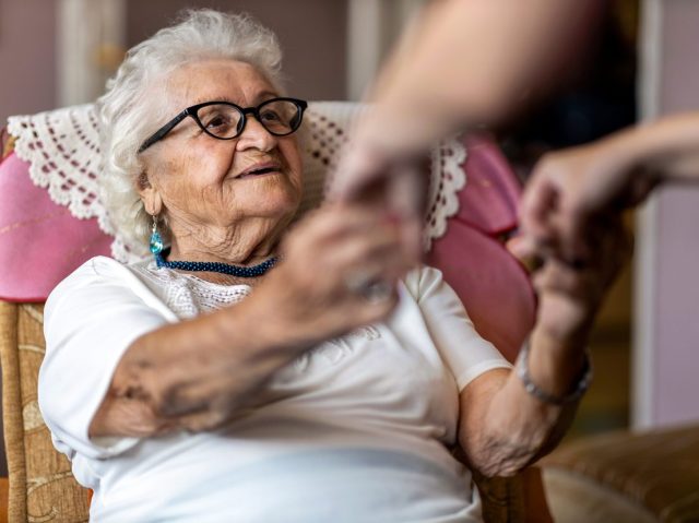 Female home carer supporting old woman to stand up from the armchair at care home